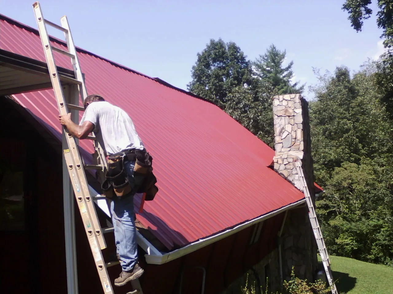 Roofer on a ladder beside a red metal roof and stone chimney — Soffit & Fascia Repair by Roofing Brothers in Glen Mills